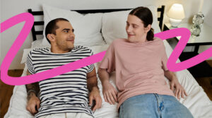 Couple lying on a bed facing each other and smiling, with a bright pink decorative swoosh across the scene.