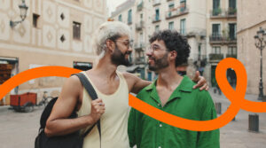 Two men with beards and glasses smile as they chat in a European street; one wears a yellow tank top and backpack, the other a green shirt, with an orange decorative ribbon across the scene.