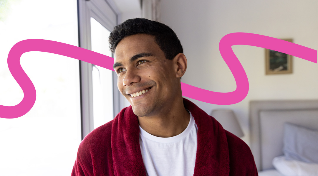 Smiling man in a white shirt and red robe stands by a sunny window with a pink abstract swirl in the background.