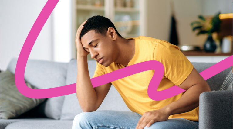 Young man in a yellow shirt sits on a sofa, head in his hand, appearing distressed or tired in a living room setting.